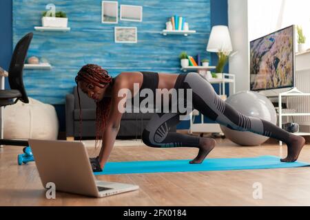 Black FIT Athletic Woman Training for muscle strength doing Mountain Climbers Positionieren Sie auf einer Yogamatte, die in Sportbekleidung-Leggings gekleidet ist, im Wohnzimmer nach Online-Anweisungen. Stockfoto