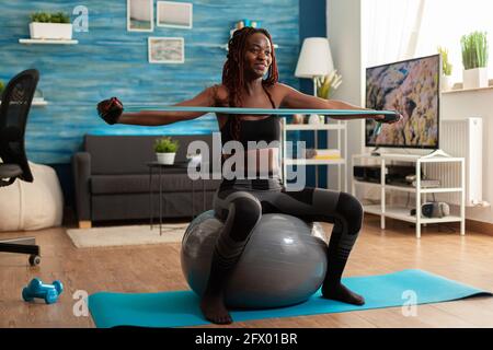 Athletische Passform fröhliche schwarze Frau, die mit Gummiband Rückenmuskulatur trainiert und auf einem Stabilitätsball im Wohnzimmer sitzt, um einen gesunden Lebensstil zu gewährleisten. Training in Sportbekleidung Leggings. Stockfoto