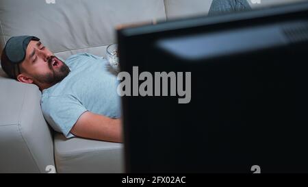 Mann mit Augenschlafmaske, der auf dem Sofa liegt, während er einen Unterhaltungsfilm mit einer Popcornschüssel in den Händen hält. Kaukasischer Mann, der während der Fernsehshow spät in der Nacht in der Küche vor dem Fernseher sitzt Stockfoto