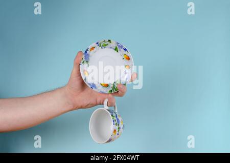 Hand und Tasse auf blauem Hintergrund. Mann hält weiße Tasse mit bemalten Blumen. Tasse aus Keramik und Untertasse für Tee oder Kaffee hell in Blau A dekoriert Stockfoto