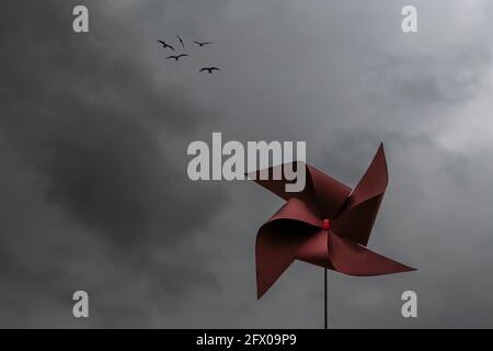 Ein rotes Windrad gegen einen dramatischen Himmel mit dunklen Wolken Und eine Vogelschar, die hoch am Himmel fliegt Stockfoto