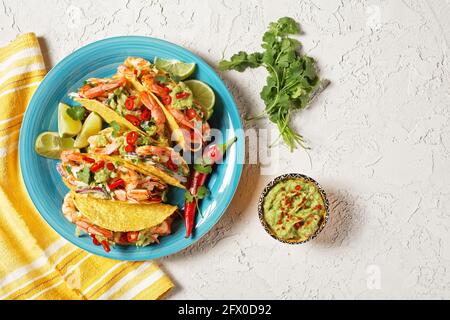 taco-Muscheln mit gegrillten Garnelen, Kohlsalat, Limette und Guacamole auf einem blauen Teller auf einem weißen Tisch, flach liegend, freier Platz Stockfoto