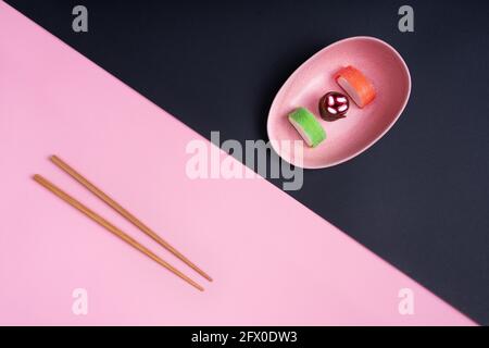 Blick von oben auf appetitlich buntes, süßes Sushi, serviert auf Keramik Teller mit Holzstäbchen auf rosa und schwarzem Hintergrund Stockfoto