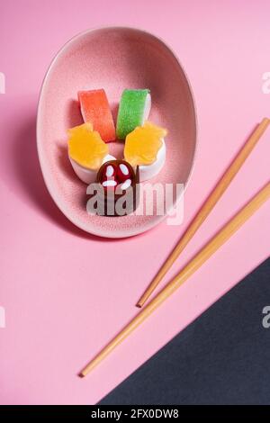 Blick von oben auf appetitlich buntes, süßes Sushi, serviert auf Keramik Teller mit Holzstäbchen auf rosa und schwarzem Hintergrund Stockfoto