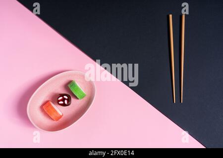 Blick von oben auf appetitlich buntes, süßes Sushi, serviert auf Keramik Teller mit Holzstäbchen auf rosa und schwarzem Hintergrund Stockfoto