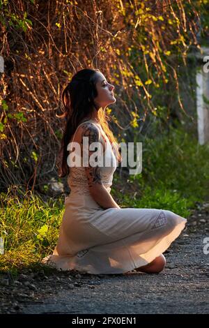 Träumende Dame mit tätowierten Arm in weißem Kleid und sitzend Auf grünem Rasen in der Natur Stockfoto