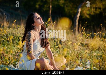 Träumende Dame mit tätowierten Arm in weißem Kleid und sitzend Auf grünem Rasen in der Natur Stockfoto
