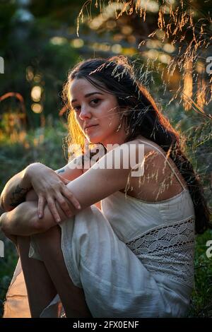 Träumende Dame mit tätowierten Arm in weißem Kleid und sitzend Auf grünem Rasen in der Natur Stockfoto
