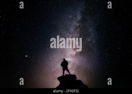 Niedriger Winkel der Silhouette des anonymen Touristen stehend mit Licht Fackel auf dem Kopf auf einer Klippe gegen den glühenden Sternenhimmel Nacht Stockfoto