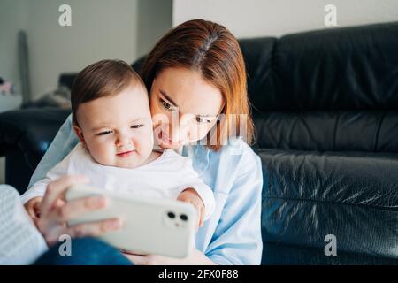 Junge Frau, die Selfie mit einem charmanten Kleinkind auf dem Mobiltelefon nimmt, während sie Zeit zu Hause verbringt Stockfoto