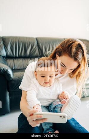 Junge Frau, die Selfie mit einem charmanten Kleinkind auf dem Mobiltelefon nimmt, während sie Zeit zu Hause verbringt Stockfoto