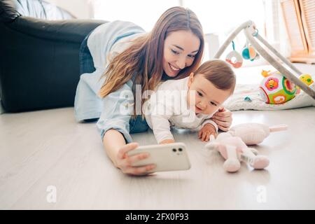 Verspielte Frau, die mit einem charmanten Kleinkind ein Selfie auf dem Mobiltelefon nimmt, während sie Zeit zu Hause verbringt Stockfoto