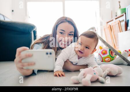 Verspielte Frau, die mit einem charmanten Kleinkind ein Selfie auf dem Mobiltelefon nimmt, während sie Zeit zu Hause verbringt Stockfoto