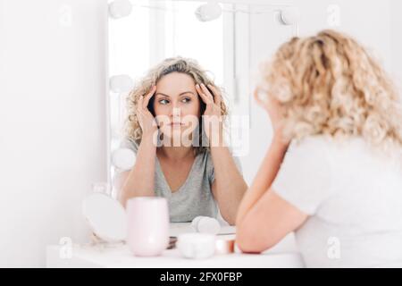 Unemotional attraktive weibliche in Casual Shirt berühren Gesicht und suchen Bei der Reflexion im Spiegel aufmerksam beim Sitzen am Eiteltisch Im Schlafzimmer Stockfoto