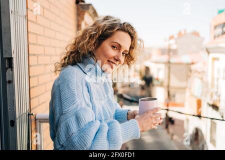 Seitenansicht einer zufriedenen jungen Frau mit lockigen Haaren in einem bequemen Kriegspullover, die am sonnigen Morgen eine Tasse heißen Kaffee trinkt, während sie sich auf dem Balkon entspannt Stockfoto