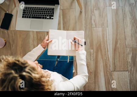 Von oben der Ernte anonyme Studentin mit lockigem Haar In legerer Kleidung am Glastisch sitzen und Notizen machen Im Copybook während der Vorbereitung auf Stockfoto