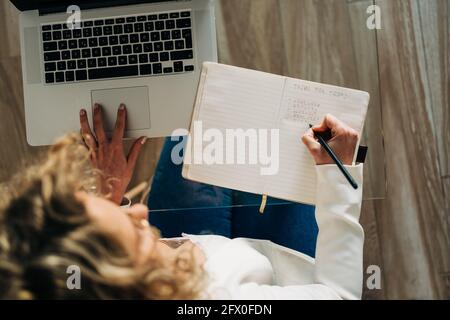 Von oben der Ernte anonyme Studentin mit lockigem Haar In legerer Kleidung am Glastisch sitzen und Notizen machen Im Copybook während der Vorbereitung auf Stockfoto