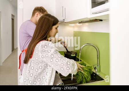 Seitenansicht einer jungen ethnischen Dame mit langen dunklen Haaren In trendiger Bluse waschen frischen grünen Salat im Waschbecken in der Nähe Anonymer Freund bereitet Essen in Sau zu Stockfoto