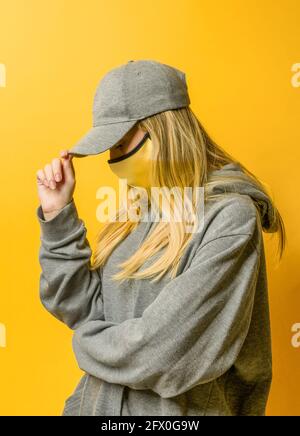 Unemotionale junge Hündin im lockeren Hoodie und gelber Gesichtsmaske Aufsetzen der Kappe und Blick auf die gelbe Wand Stockfoto