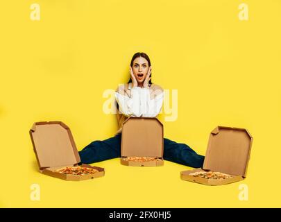 Emotional überrascht junge Frau von drei Pizzen in Boxen auf gelbem Hintergrund umgeben. Pizzalieferkonzept. Stockfoto