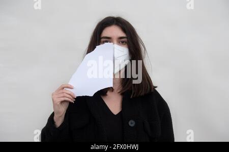 Frau in Corona-Pandemie mit FFP2-Maske hält ein zerrissenes Stück Papierhälfte vor ihr Gesicht, um eine Nachricht oder Informationen einzufügen, Leerzeichen zu kopieren, Stockfoto