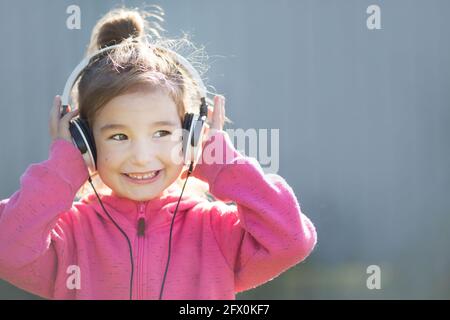 Ein fröhliches Mädchen mit Kopfhörern und einem pinken Hoodie hört Musik und lacht. Draußen, grauer Hintergrund Stockfoto