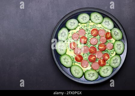 Omelette mit Kirschtomaten, grünen Zwiebeln und Würstchen auf einem Teller auf dunklem Grund. Draufsicht, flach liegend Stockfoto