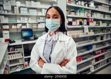 Glückliche weibliche Frau, die in der Apotheke arbeitet und hinter dem Tresen steht Labcoat und Covid-19 Gesichtsschutzmaske Stockfoto