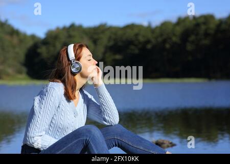 Seitenansicht Porträt einer entspannten Frau, die Musik hört Mit kabellosen Kopfhörern, die in einer Lagune in den Bergen sitzen Stockfoto