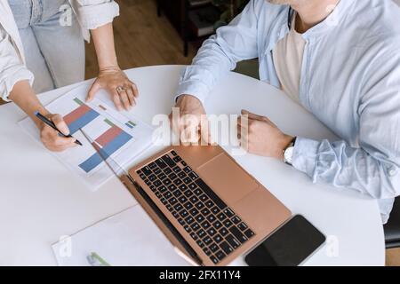 Kollegen sitzen am Arbeitsplatz im modernen Co-Working-Bereich und diskutieren mit einem Laptop Online-Finanzdiagramme und führen angenehme, informelle Gespräche Stockfoto