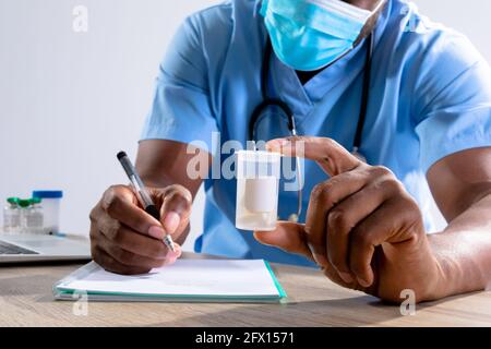 afroamerikanischer männlicher Gesundheitshelfer trägt Gesichtsmaske mit Medikamenten Behälter Notizen machen Stockfoto