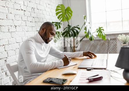 Serious Millennial African American Geschäftsmann sitzen am Schreibtisch im Home Office Arbeit online auf Laptop mit Dokumenten. Nachdenklicher Mann schaut sich das Notizbuch an Stockfoto