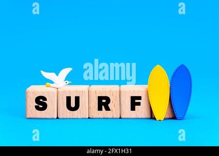 Word SURF aus hölzernen Alphabet-Blöcken mit zwei Surfbrettern und einer Spielzeugmöwe auf blauem Hintergrund. Stockfoto