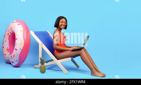 Online arbeiten vom tropischen Paradies aus. Schwarze Frau im Bikini, die auf einem Loungesessel sitzt und einen Laptop auf blauem Hintergrund verwendet Stockfoto