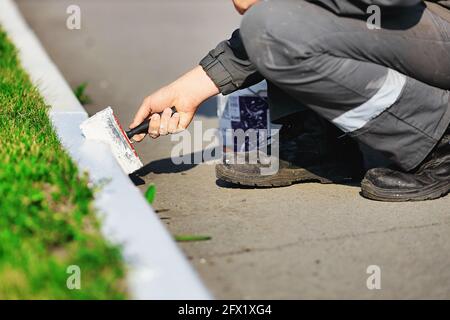 Ein Arbeiter in Overalls malt an einem Sommertag eine Grenze mit weißer Farbe. Städtische Dienstleistungen, Landschaftsbau. Die Hand hält einen Pinsel. Nahaufnahme Stockfoto