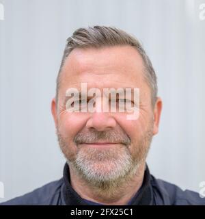Glücklicher Mann mittleren Alters, der mit einem Lächeln auf die Kamera blickt Als er vor einer weißen Wand in posiert Ein Porträt von Kopf und Schultern Stockfoto