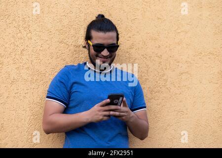 Junger Latino-Mann mit Sonnenbrille, die im Telefon SMS schreibt und lächelt. Technologiekonzept Stockfoto