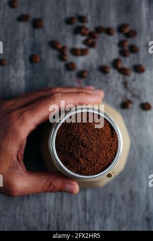 Blick aus der Perspektive eines jungen kaukasischen Mannes, der Kaffee in einer beigen Mokakapfanne zubereitet, an einem grauen rustikalen Holztisch mit einigen gerösteten Kaffeebohnen bestreut Stockfoto