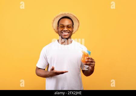 Millennial schwarzer Kerl zeigt Glas leckeren tropischen Cocktail auf Gelber Studiohintergrund Stockfoto