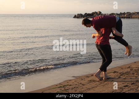 Zwei junge kaukasische Männer spielen bei Sonnenuntergang am Strand. Ein Mann, der seinen Freund auf der Schulter trägt. Lustiger Sommer-Lifestyle, Freundschaft und Brüderlichkeit. Stockfoto