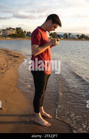 Junger kaukasischer Mann, der bei Sonnenuntergang am Strand mit Handy steht. Rotes T-Shirt. Denia, Alicante, Spanien. Mediterraner Abend. Stockfoto