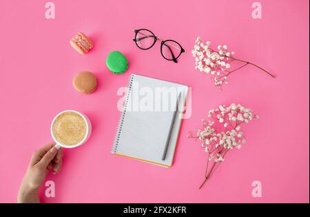 Nachbau für Prüfliste. Weibliche Hand hält Tasse Kaffee mit Kuchen Makronen, saubere Notebook, Gläser und Blumen auf dem Tisch Stockfoto