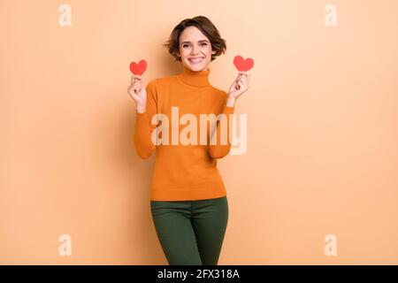 Foto von süßen liebenswert junge Mädchen orange Rollkragen hält zwei Rote Herzen isoliert beige Farbe Hintergrund Stockfoto