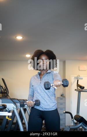 Ernsthafte Frau, die mit Hanteln im Fitnessstudio zu Hause trainiert. Nahaufnahme, eine Person, Fitnessmodell, afroamerikanisch, afrikanisches Modell, Sportbekleidung, Naturhaar m Stockfoto