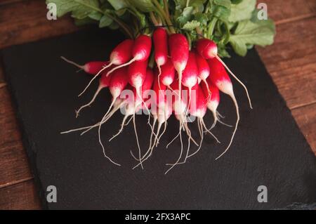 Frischer Rettich mit Stielen und Blättern liegt auf einem schwarzen Podest-Brett aus Granit Stockfoto