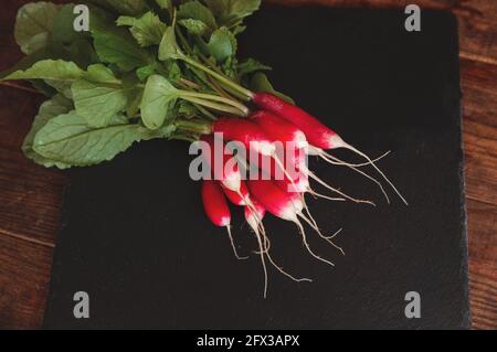 Frischer Rettich mit Stielen und Blättern liegt auf einem schwarzen Podest-Brett aus Granit Stockfoto