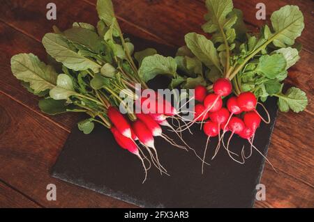 Frischer Rettich mit Stielen und Blättern liegt auf einem schwarzen Podest-Brett aus Granit Stockfoto