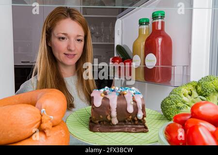 Offener Kühlschrank von innen, Glasregale mit verschiedenen Lebensmitteln. Ungesunde Ernährung, Zucker Lebensmittel Konzept. Junge Frau, die Schokoladenkuchen anschaut Stockfoto