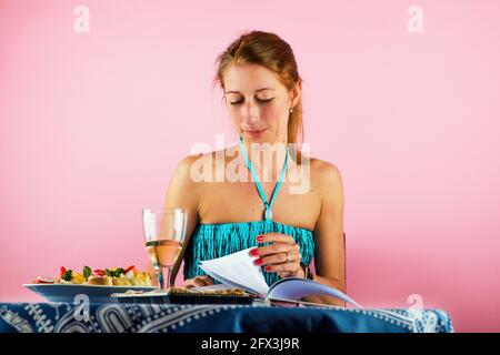 Junge attraktive kaukasische alleinerziehende Frau sitzt bei einem Essen und einem Glas Wein am Tisch, liest Buch und wendet sich um. Auf rosa pastellfarbenem Hintergrund. Stockfoto