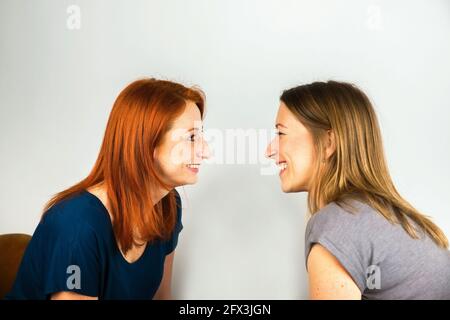 Zwei attraktive junge Frauen lachen sich gegenseitig aus. Sie sitzen einander gegenüber und haben Spaß. Auf grauem Hintergrund. Stockfoto